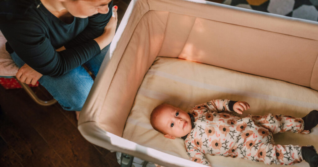 A baby in a bassinet with a woman sitting down next to them and smiling at them