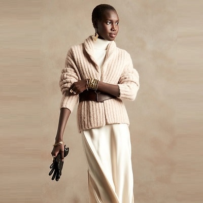 A Black woman with very short hair wearing a cream colored shirt, cream colored flowy pants, and a darker cream colored sweater. She is holding black gloves.