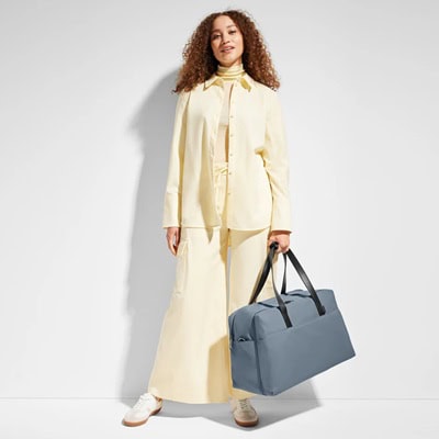 A woman wearing an all pale yellow outfit and carrying a dark gray Away tote 