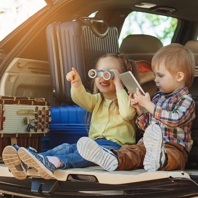 Two young blonde children in the open hatchback of a car with suitcases behind them; the girl is looking through binoculars and the boy is looking at a book