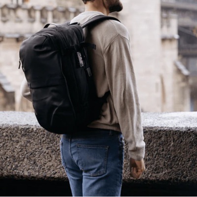 A man on a stone balcony wearing a gray shirt, blue jeans, and a black travel bag on his shoulders
