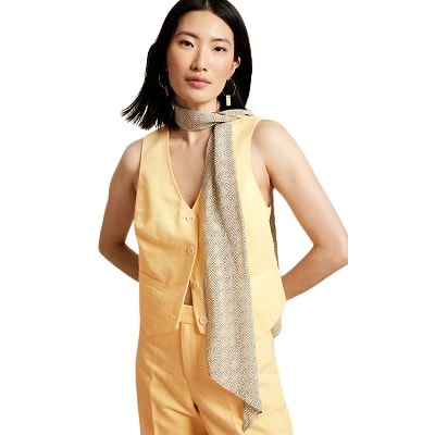 A woman with shoulder-length black hair wearing a yellow vest with matching tailored pants and a white-and-beige geometric-print scarf