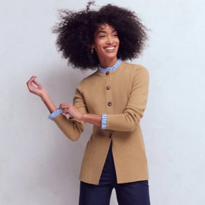 A woman wearing a camel-colored collarless jacket, light blue blouse with ruffles, and navy pants