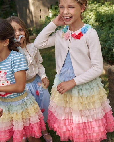 Three girls wearing colorful Boden clothing; the main girl is wearing an off-white cardigan with flower appliques over a rainbow tulle dress