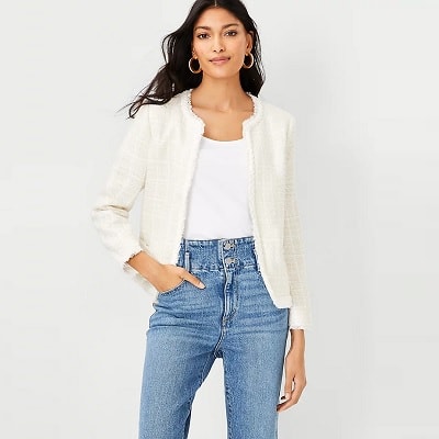 A woman with long black hair and hoop earrings wearing a cream-colored tweed jacket, white top, and medium-blue jeans