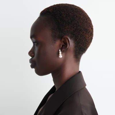 A woman in profile view; she is wearing stud earrings with four pearls each, and a brown blazer
