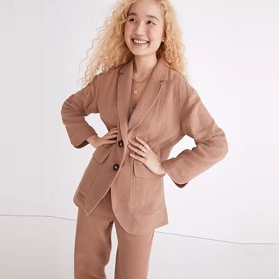 A smiling woman with long, blonde, wavy hair wearing a light brown blazer, light brown pants, and two small gold necklaces