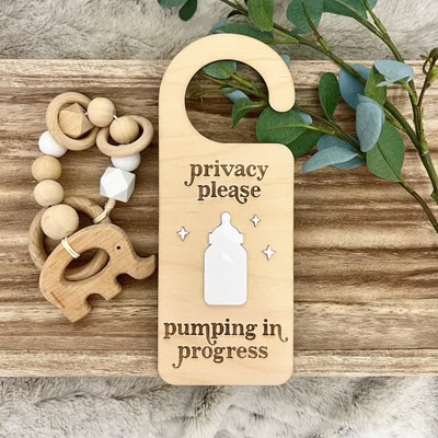 A wooden "pumping in progress" sign with a bottle on it. The sign is next to a leafy stalk and elephant wooden baby toy. All are on a board on a fuzzy gray blanket