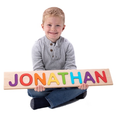 A boy wearing a gray sweater and blue jeans, holding a wooden puzzle with the letters "Jonathan" in multiple colors