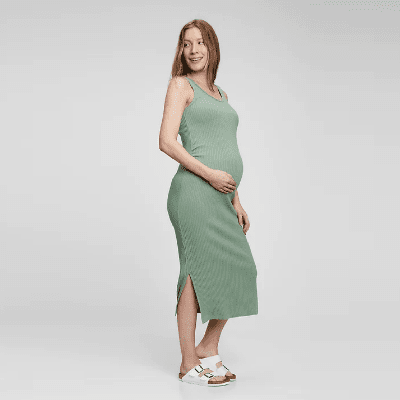 A pregnant woman with long brown hair wearing a green maternity dress and white sandals
