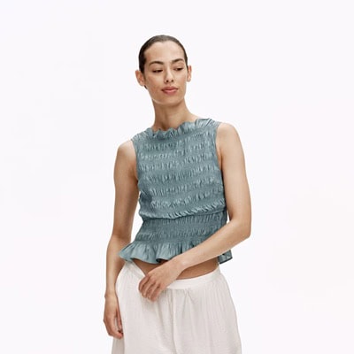 A woman wearing a light blue smocked, sleeveless nursing top and flowy white pants