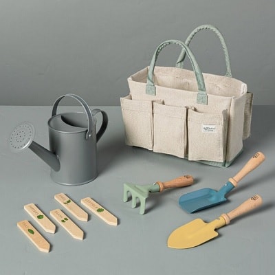 A canvas tote bag next to a gray watering can, plant stakes, and three gardening tools; all are sized for kids