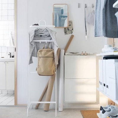 A room containing a white rack holding a shirt and backpack, a mirror, a wall clock, white shoe cabinets, two pairs of shoes, and a brown floor mat