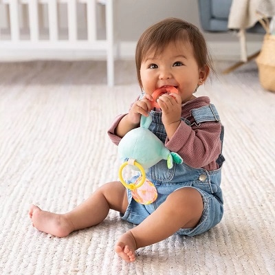 A baby in a purple shirt and shortalls in front of a crib, chewing on a toy