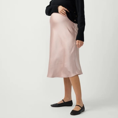 A pregnant woman wearing black, flats, a black top, and a light pink satin midi skirt
