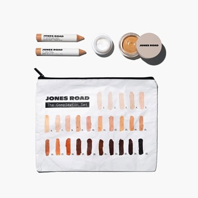A makeup bag reading "Jones Road The Complexion Set" and depicting many shade swatches, next to two makeup pencils and and eye cream and foundation containers