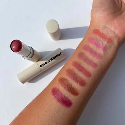 An arm with eight different pink swatches of Jones Road Lip and Cheek Stick, next to a the product and an empty container 

