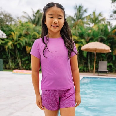 A girl wearing a purple rash guard and purple print swim shorts. She is standing in front of an outdoor pool.