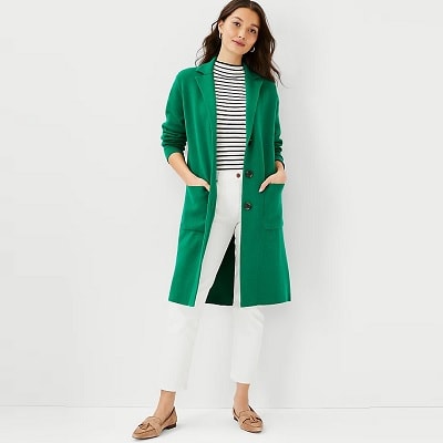 A woman with medium skin tone and long brown hair wearing a long green coat, striped shirt, white pants, and loafers