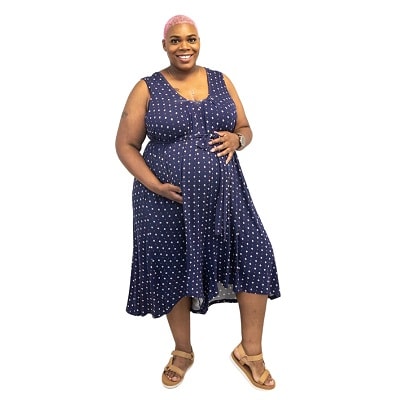 A pregnant woman with close-cropped pink hair wearing a blue nursing romper with white polka dots, with light brown sandals