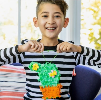 A boy with short black hair and a black-and-white striped shirt holding up a handmade piñata in the shape of a potted cactus 