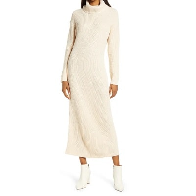 Woman with head cropped out of photo wearing a long, cream-colored sweater dress and white short boots