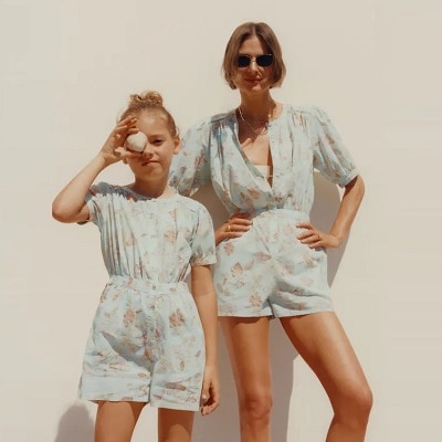 A woman and young girl wearing similar floral rompers. The woman is wearing sunglasses and the girl is holding a seashell in front of her eye