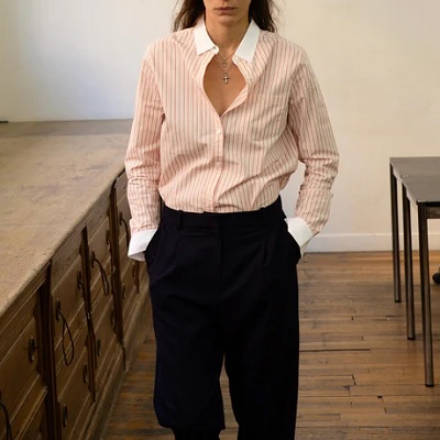 A woman wearing a pink-and-beige pinstripe blouse with black pants; she is sitting on a large wooden piece of furniture.