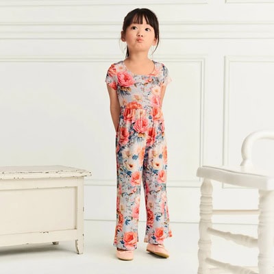 A girl wearing a floral jumpsuit with pink, blue, and orange elements. She is standing next to a white bench and white chair in front of a white paneled wall.