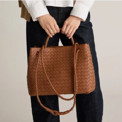 A brown leather handwoven satchel bag held by a woman wearing a white top and black pants