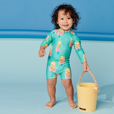 A toddler with curly black hair wearing a rashguard swimsuit with a popsicle print, carrying a yellow bucket