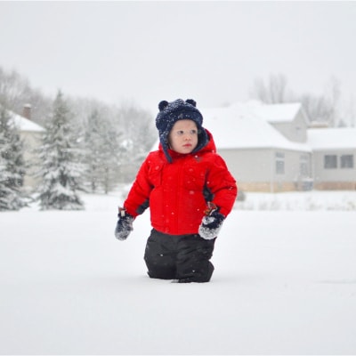 small boy wears a bright red coat and blue bear hat, standing in a suburban snow drift with trees and houses behind him