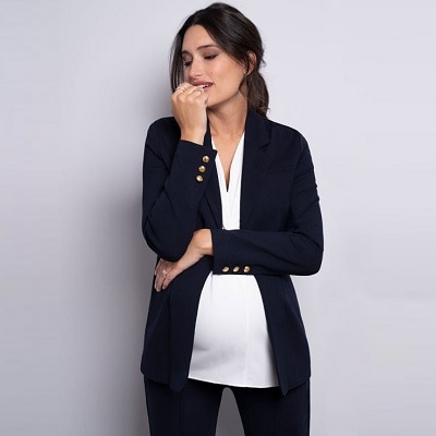 A pregnant woman wearing a navy maternity blazer with gold buttons on both cuffs, with a white top and navy pants
