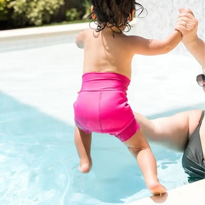 A toddler wearing a pink reusable swim diaper, jumping into a pool while holding hands with an adult whose body is cropped out
