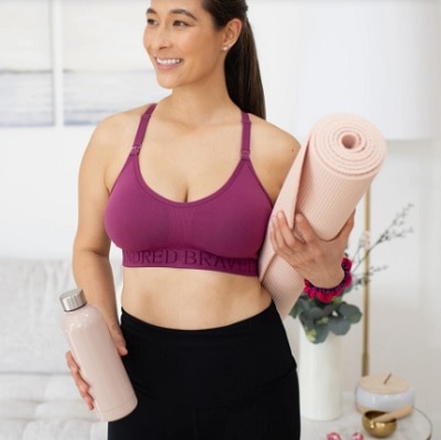 A woman wearing a purple nursing sports bra and black pants, holding a water bottle and yoga mat