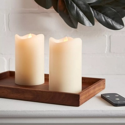 Two cream-colored LED candles on a wooden tray on a shelf with a remote
