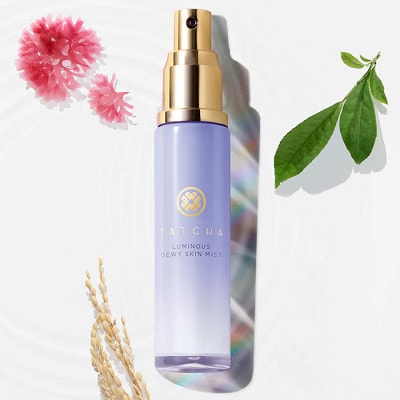 A purple-and-gold spray bottle of Tatcha Luminous Dewy Skin Mist on a white background, next to small pink flowers, green tea leaves, and a rice stalk