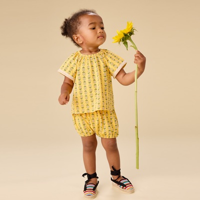 A young girl wearing a matching yellow floral shorts-and-shirt set and multicolored striped shoes. She is holding a sunflower and looking at it.
