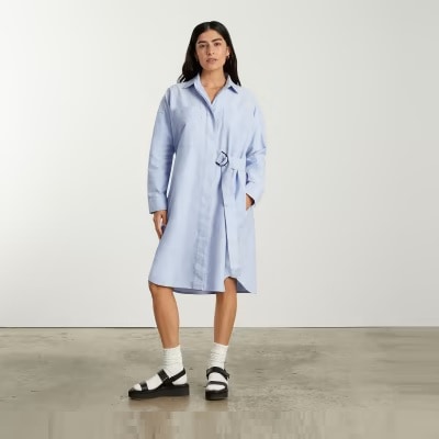 A woman with long black hair wearing a light blue shirtdress with white socks and black sandals