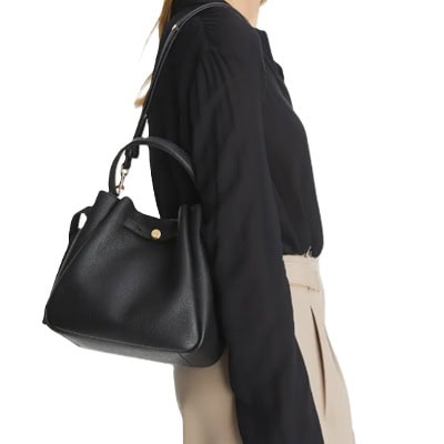 A woman wearing a black top and khaki-colored pants, carrying a black bucket bag
