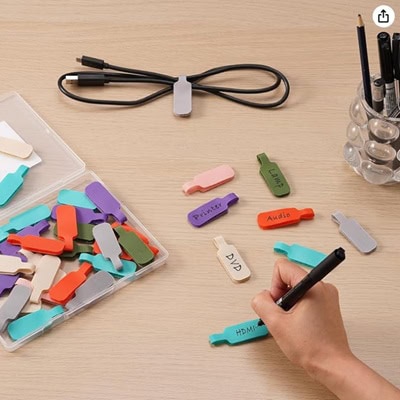 The image shows a clear box of colorful small cable labels, next to a person's hand labeling one. There is a gray-labeled black cable next to a clear container of black pens.