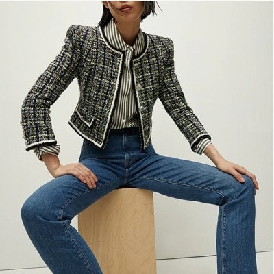 A woman wearing a tweed jacket, striped blouse, and jeans, sitting on a wooden stool