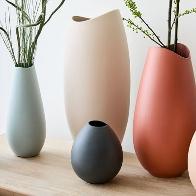 Four ceramic vases -- light blue, cream, dark gray, and orange -- on a wooden table. Two of them have greenery in them.