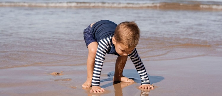 baby plays on beach by putting hands in wet sand; he wears a stripey rashguard. 