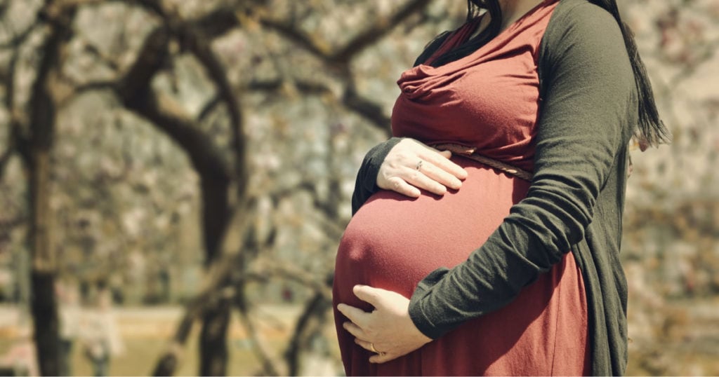 professional young woman holds her very pregnant belly; she's wearing a coral dress, army green sweater and standing in the woods. 