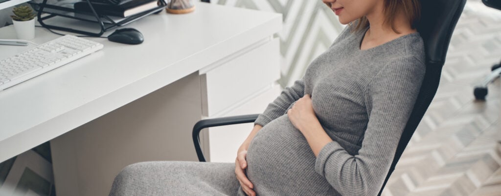 professional woman relaxes in chair at the office; she is pregnant and wearing a gray maternity dress