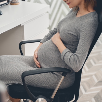 professional woman relaxes in chair at the office; she is pregnant and wearing a gray maternity dress