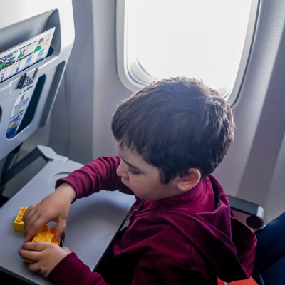 little boy plays with yellow Legos on a gray airplane tray; he is sitting in front of an airplane window and his parents have mastered carry-on only travel with kids