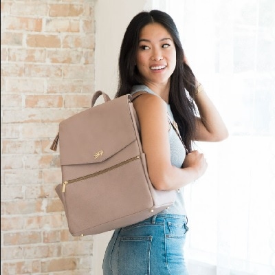 An Asian woman with long black hair wearing a gray tank top and jeans and carrying a tan backpack that is a diaper bag (legs cropped out). She is in front of a brick wall.