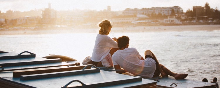 a woman and a man relax on a boat or pier, overlooking water, with buildings in the distance; the light suggests sunset. 